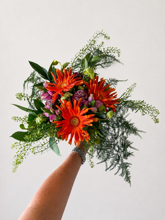 A hand holding a vibrant floral bouquet featuring bright orange gerbera daisies, green foliage, and small purple flowers against a plain light gray background.