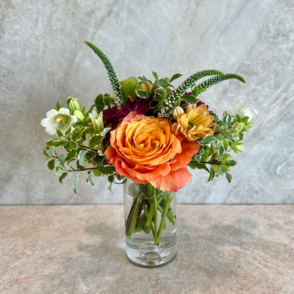 A vibrant floral arrangement featuring a prominent orange rose, yellow ranunculus, white snowdrops, and green foliage, presented in a clear glass vase on a stone-tiled surface against a textured gray wall.