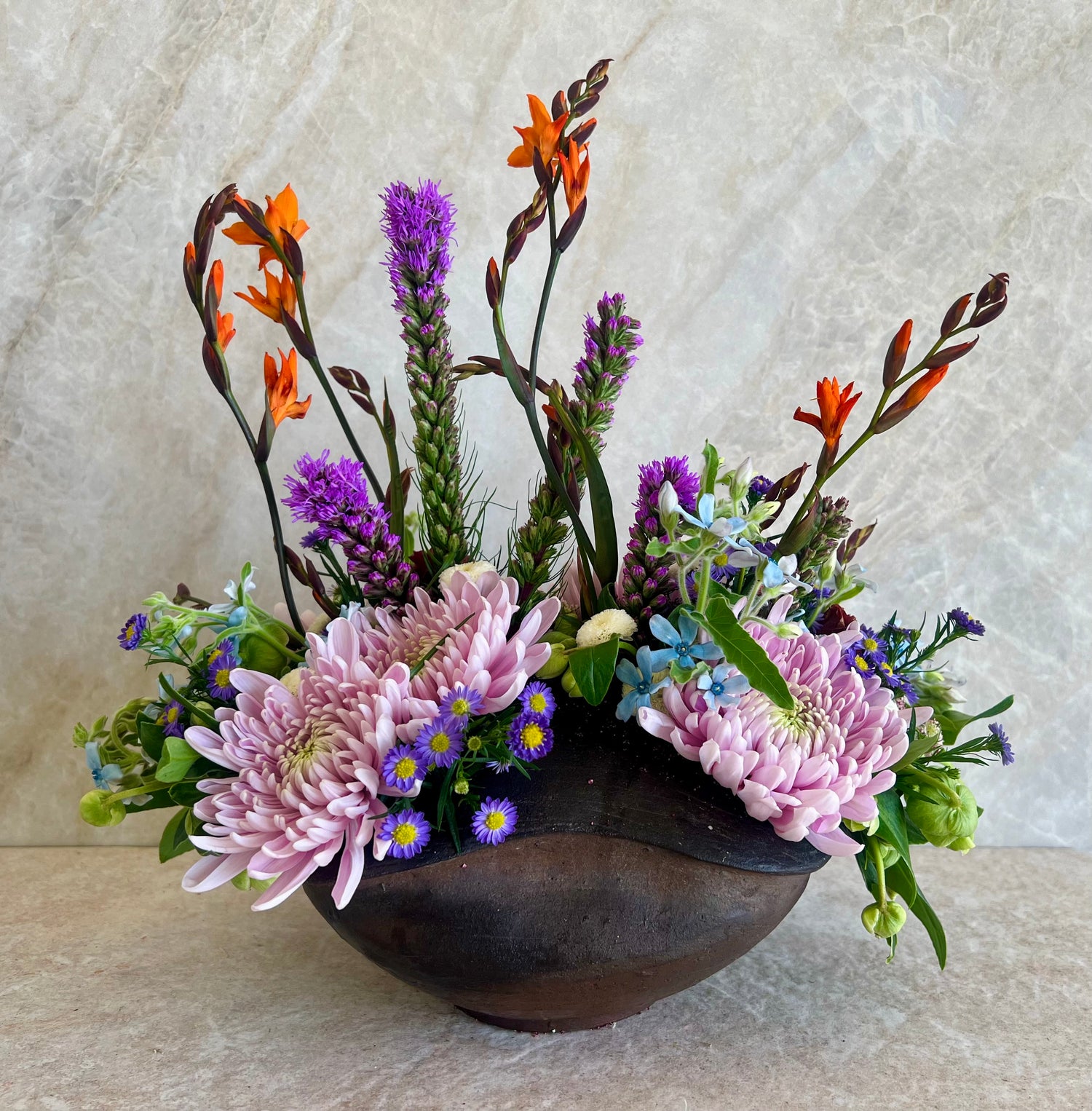 A floral arrangement featuring light pink chrysanthemums, purple spikes, orange flowers, and small blue blossoms in a dark brown ceramic bowl, set against a textured beige background.