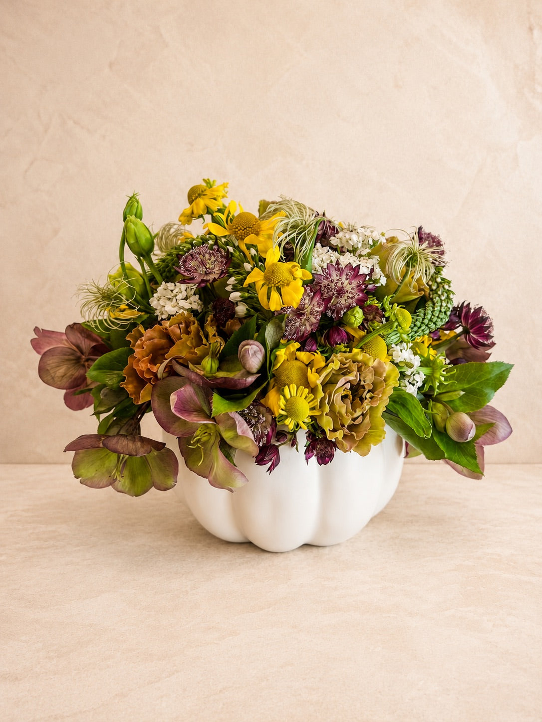 Arrangement of flowers in yellows, purple, pinks, greens, and white within a white ceramic pumpkin vase.